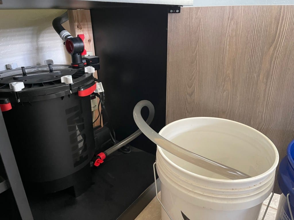 Filtro externo de acuario bajo un mueble, con la manguera vaciándose en un cubo durante la limpieza